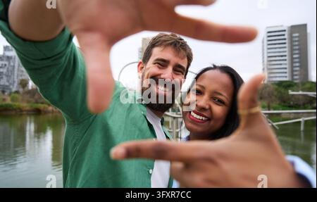 Happy multiracial couple taking a photo framing with hands in a city park Stock Photo