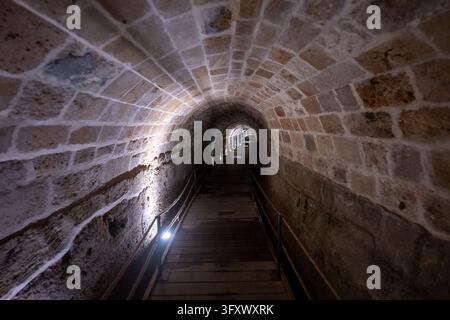 Stone-vaulted Templar Tunnel in Acre, Israel, with wooden walkway, metal railings, and low accent lighting along ancient limestone walls. Stock Photo