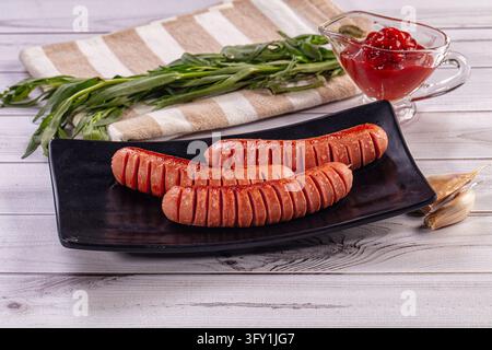 Three browned sausages served with dipping sauce, garlic cloves, and fresh herbs on striped linen. Savory meat platter Stock Photo