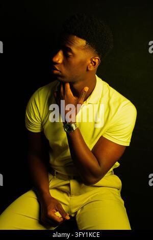 Young man in yellow clothing, sitting thoughtfully with a hand on his chin in dramatic light Stock Photo