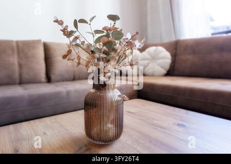 Cozy living room interior featuring ribbed glass vase with dried eucalyptus branches on wooden coffee table with brown sofa in background Stock Photo