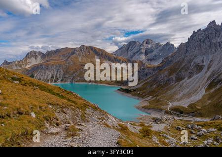 Hiker explores tranquil turquoise lake surrounded by rugged autumn-hued alpine peaks under dynamic cloud formations evoking awe and peace Stock Photo