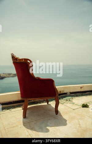 An antique red velvet armchair stands on a sunlit seaside terrace, casting a long shadow across pale stone tiles with the calm sea behind it. Stock Photo