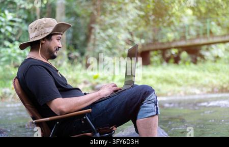 Happy adult handsome Asian man sitting on camping chair at stream in the forest and working on laptop. Workation concept. Intrnet 5G Stock Photo