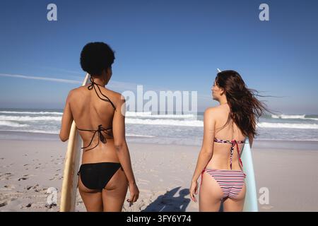 Rear view of young multi-ethnic women holding their surfboards while standing in front of the sea and looking at the waves on the beach in the sunshin Stock Photo