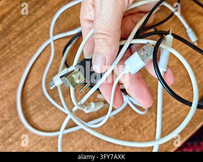 A hand holds a bundle of tangled white and black charging cables with connectors and adapters over a wooden tabletop, illustrating everyday electronic Stock Photo