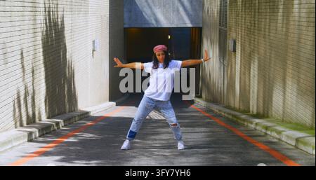 A young African American woman is performing a dance move in a sunlit alley, with copy space. Her casual attire and dynamic pose suggest a moment of c Stock Photo