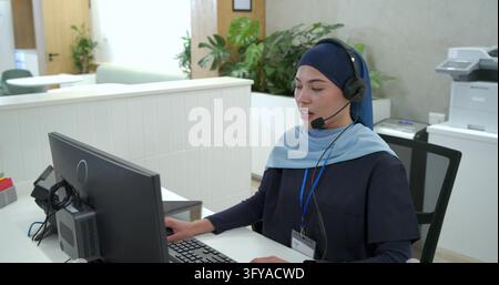 Female receptionist sitting at front desk wearing navy hijab and headset, using mouse, copy space. Office, reception, monitor, keyboard, plants, dayli Stock Photo