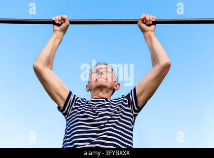 Portrait of an elderly man wearing glasses doing exercises on a horizontal bar at a sports ground. Staying active after retirement. Stock Photo