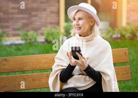 Happy older woman checks smartphone while sitting on wooden bench outdoors. Calm woman enjoys garden leisure in elegant white clothing, smiling Stock Photo