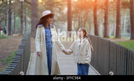 Mother and daughter stroll outdoors together along wooden bridge walkway in park, holding hands while smiling warmly at each other amidst peaceful Stock Photo