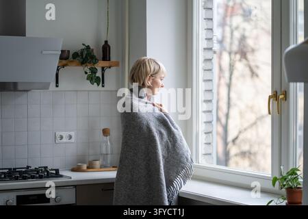 Thoughtful woman wrapped in blanket looks through kitchen window on cold morning without heating Stock Photo