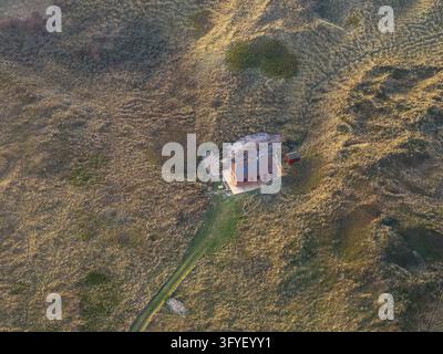 Aerial view of a small house nestled in the sandy dunes of Vejlby Klit, surrounded by grass and a dirt path in Thyboron, Denmark. Stock Photo