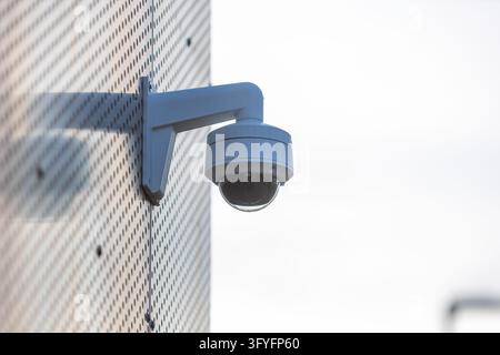 A white dome security camera is securely mounted on the exterior wall of a modern building featuring a distinctive perforated metal facade, capturing Stock Photo