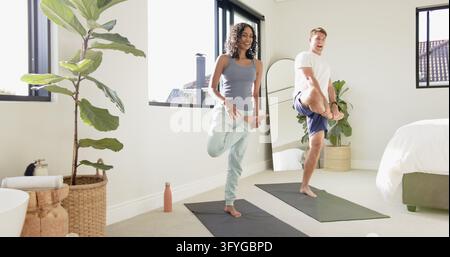 Diverse couple practicing yoga pose on gray yoga mats in bedroom with potted plant, water bottle. Wellness, fitness, relaxation, balance, home studio, Stock Photo