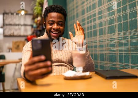 Smiling young man waving during a video call on his smartphone while sitting at a cafe table. Stock Photo