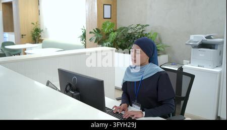 Typing receptionist wearing navy hijab and lanyard, entering data at reception, monitor, copy space. Office, modern, bright, professional, computer, h Stock Photo