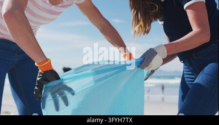 Two diverse female friends putting rubbish in refuse sacks at the beach. eco conservation volunteers, beach clean-up. Stock Photo