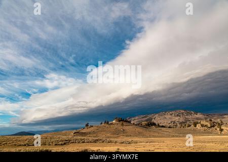 A dramatic atmospheric photograph capturing a dark storm front rolling over the expansive grassy landscape of Lamar Valley in Yellowstone National Park, USA. Dark, heavy rain clouds sweep across the sky, casting deep shadows over the rolling hills and sagebrush plains of the Northern Range, whilst golden late-afternoon sunlight illuminates portions of the valley floor in stark contrast. The vast open vista highlights the wild, untamed natural beauty and dynamic weather patterns characteristic of this An optimised version of a US National Park Service Image. Credit: NPS / J Frank Stock Photo