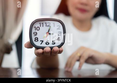 Mature woman hand holding black and white alarm clock at brown wooden table on light room background. Point of view shot. Concept of female biological Stock Photo