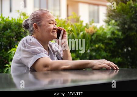 Side view portrait of Asian senior woman talking with her friends on the smartphone while relaxing in the garden. Stock Photo