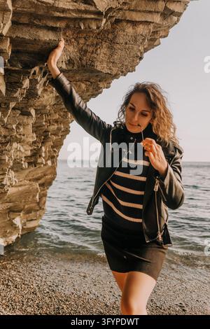 Woman jacket beach young woman wearing a leather jacket and striped sweater poses by a rocky cliff at the sea Stock Photo