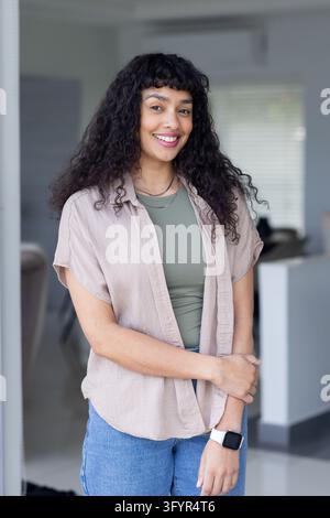 African American woman standing and smiling in bright room while wearing beige shirt and smartwatch. Portrait, confident, arms crossed, casual, profes Stock Photo
