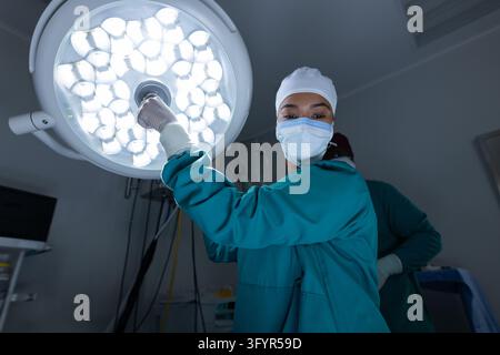 Female medical professional in teal scrubs reaching toward surgical light in operating room. Healthcare, hospital, surgery, surgeon, clinician, proced Stock Photo