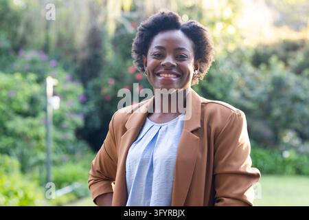 African American woman standing in garden facing camera wearing brown blazer and light blue blouse. Portrait, outdoors, nature, professional, confiden Stock Photo