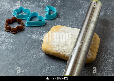 Stainless steel rolling pin lying on a block of golden shortbread cookie dough dusted with flour on a dark gray stone surface. Blue star, heart and cr Stock Photo