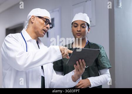 Diverse medical professionals in lab coat and scrubs reviewing tablet in hospital corridor. Healthcare, teamwork, technology, consultation, diagnosis, Stock Photo