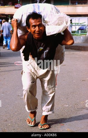 local man overladen carrying large heavy sack on his back denpasar market bali Stock Photo