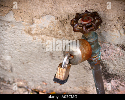 Rusted, padlocked water faucet in front of a speckled concrete wall. Stock Photo