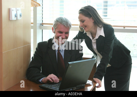 Middle aged business man working on portable computer with woman lenaing over his shoulder and looking at him. Stock Photo