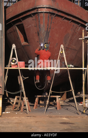 View of boat yard workshop & back view of workman sitting on trestles at work on welding repairs steel bow of boat in boatyard Bristol England UK Stock Photo