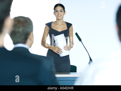 Businesswoman standing next to microphone, men blurred in foreground Stock Photo