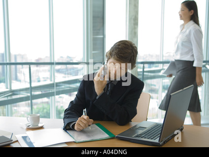 Businessman using cell phone at desk while female office worker walks by in background Stock Photo