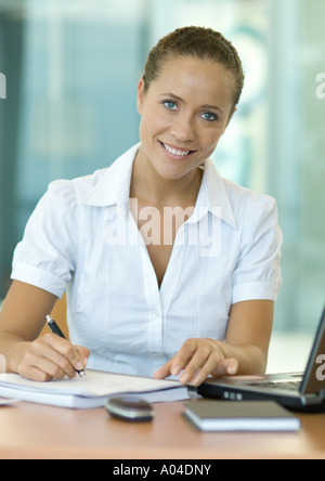 Businesswoman sitting at desk, writing, smiling at camera Stock Photo