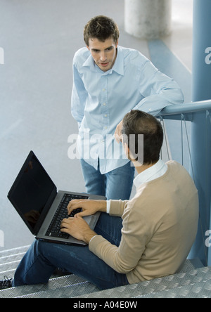Man sitting on steps, using laptop, speaking to second man Stock Photo