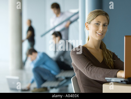 Young woman using laptop, smiling at camera, busy office in background Stock Photo