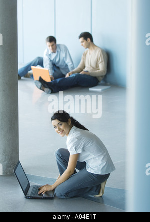 Young woman crouching sitting on floor, using laptop, men sitting in background Stock Photo