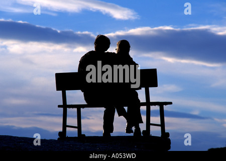 man and woman arm and arm hugging on a park bench watching the sunset in silhouette Stock Photo