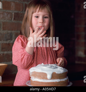 Young girl licking the icing off her fingers from a cake in front of her Stock Photo