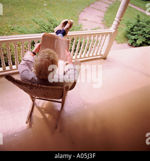 View from behind of a mature white hair man sitting on a rocking chair on the front porch reading the newspaper Stock Photo