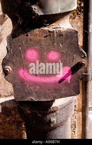 Pink sprayed Smiley sign on metal plate, weathered industrial pipe fitting with graffiti art, urban street art on old rusty metal surface. Stock Photo