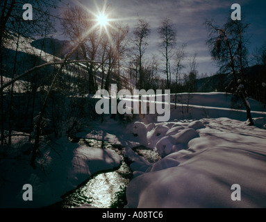 Icy freezing Winter stream flows through snow banks under a sunburst in the frosty sky Stock Photo