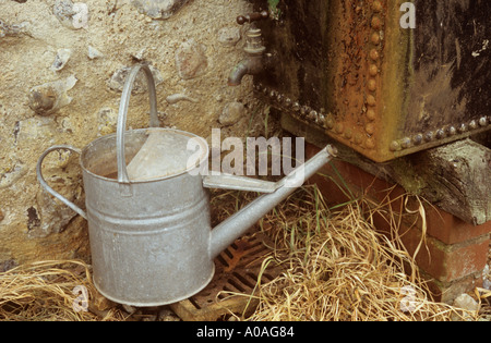 Galvanised aluminium watering can set on a drain cover below the tap of a water tank by a flint and cement wall Norfolk England Stock Photo