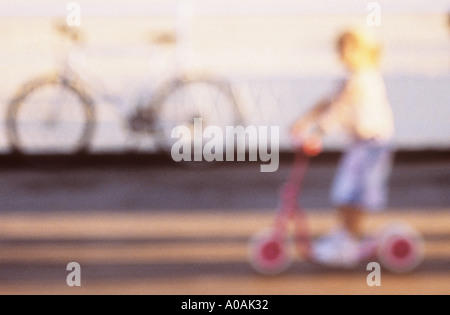 Defocussed girl on scooter whizzing past a bicycle propped against railings in summer evening sun Stock Photo