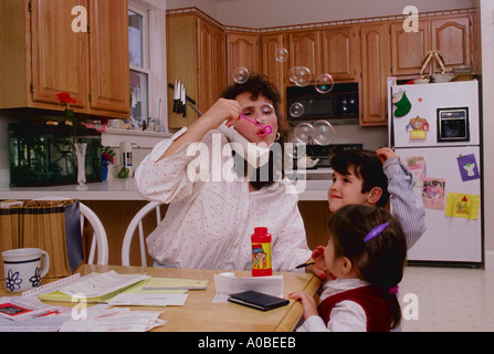 A mother talks on telephone and blows bubbles for her son and daughter at the same time Family life centers around the kitchen Stock Photo