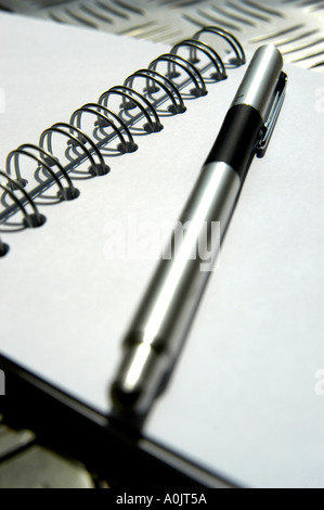White note book with black spiral binding, stainless steel pen. Shot on funky punched stainless steel table top. Stock Photo
